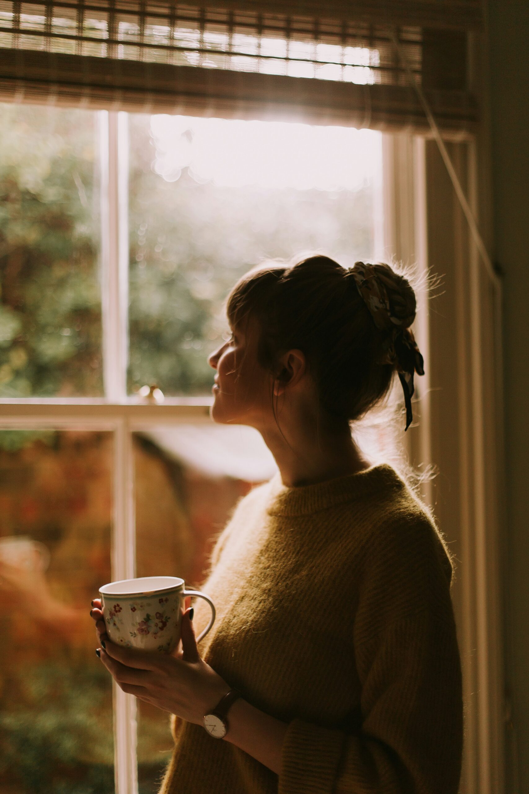 Woman holding a mug and looking out a window at home, practicing a moment of somatic awareness and nervous system regulation